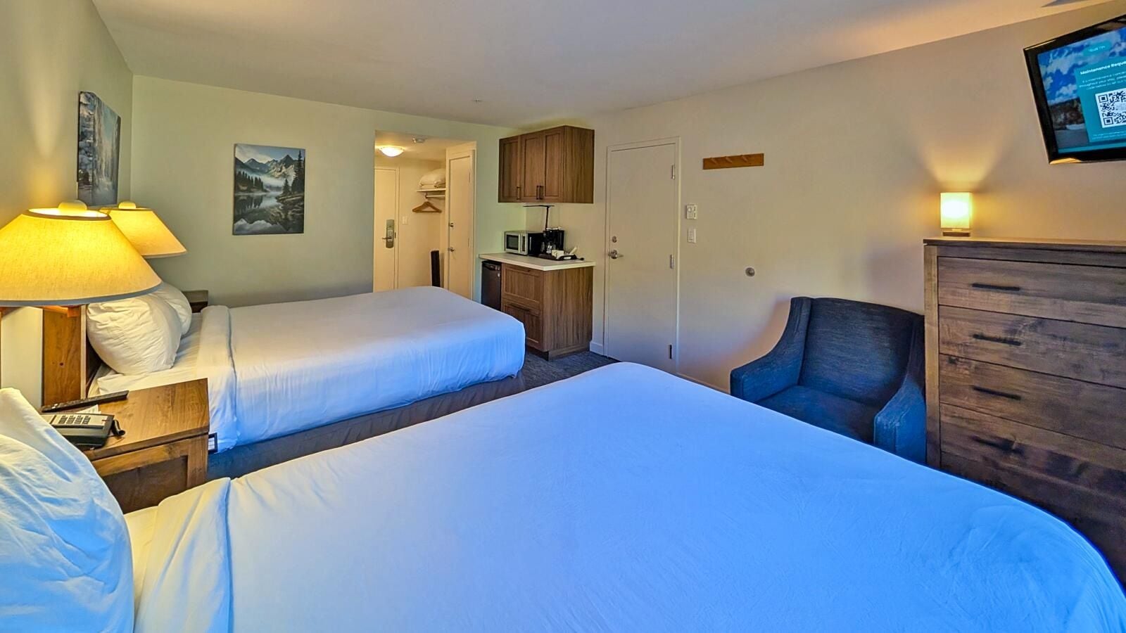 A hotel room with two beds, a desk with a lamp, a chair, a wardrobe, and a television on the wall.