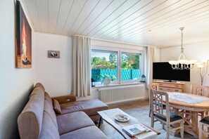 A modern living room with a sofa, dining table, chairs, a TV, and a window with a view of greenery. - Coastal Holiday Apartment (Schortens)