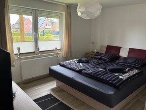 Une chambre à coucher avec un lit, une table de chevet, une lampe, une fenêtre avec des rideaux et une vue sur les maisons à l’ext érieur.