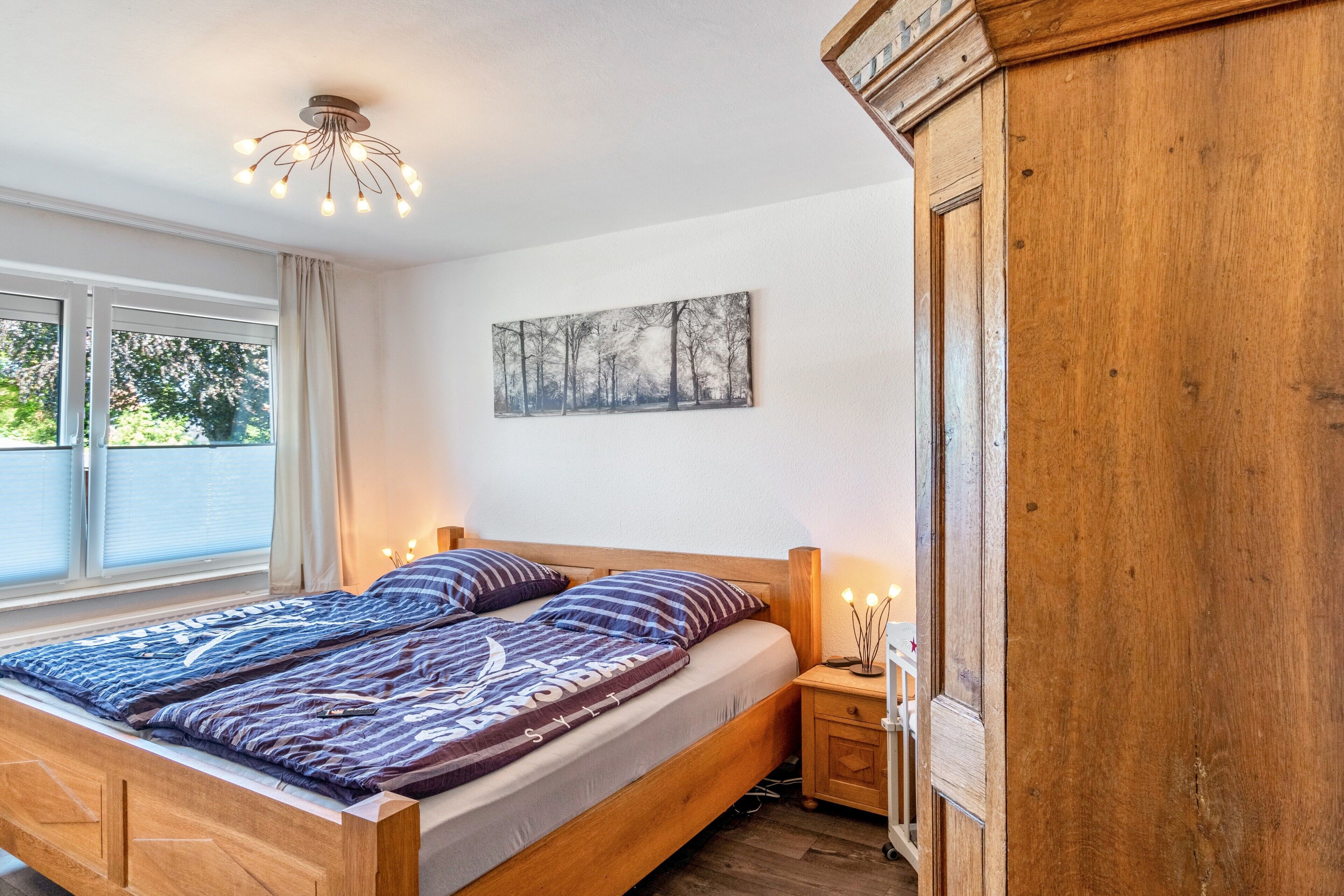 A bedroom with a wooden bed, a nightstand, a wall-mounted light fixture, and a window with curtains.