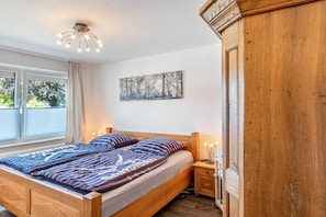 A bedroom with a wooden bed, a nightstand, a wall-mounted light fixture, and a window with curtains. - Coastal Holiday Apartment (Schortens)