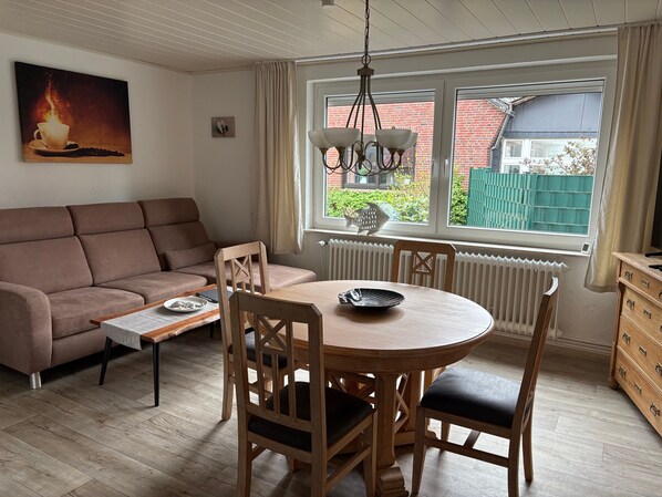 Un salon chaleureux avec un canapé, une table à manger et des chaises, un radiateur et une fenêtre donnant sur un mur de briques et de la verdure.