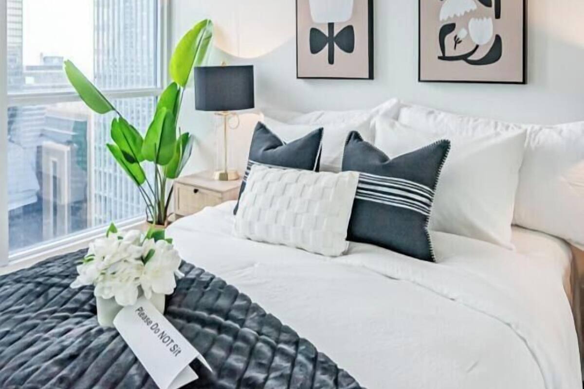 Een hotelkamer met een bed, een plant, twee kunstwerken aan de muur en een raam met uitzicht op de stad.