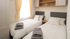 Un dormitorio con dos camas, una ventana con cortinas, una repisa de madera con un adorno en forma de corazón y una mesita de noche.