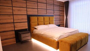 A modern bedroom with a wooden panel wall, a tufted headboard, a bedside table, a large bed with a matching ottoman, and a window with blinds.