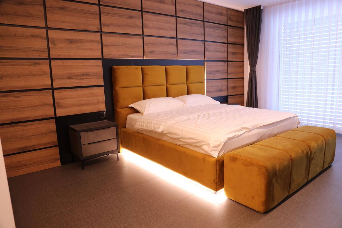 Un dormitorio moderno con pared de paneles de madera, cabecera capitonada, mesita de noche, cama grande con reposapiés a juego y una ventana con persianas.