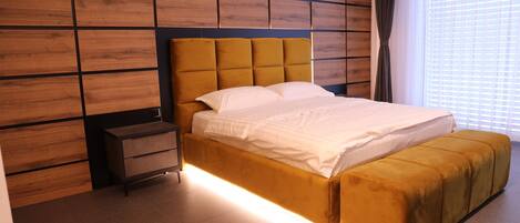 A modern bedroom with a wooden panel wall, a tufted headboard, a bedside table, a large bed with a matching ottoman, and a window with blinds.
