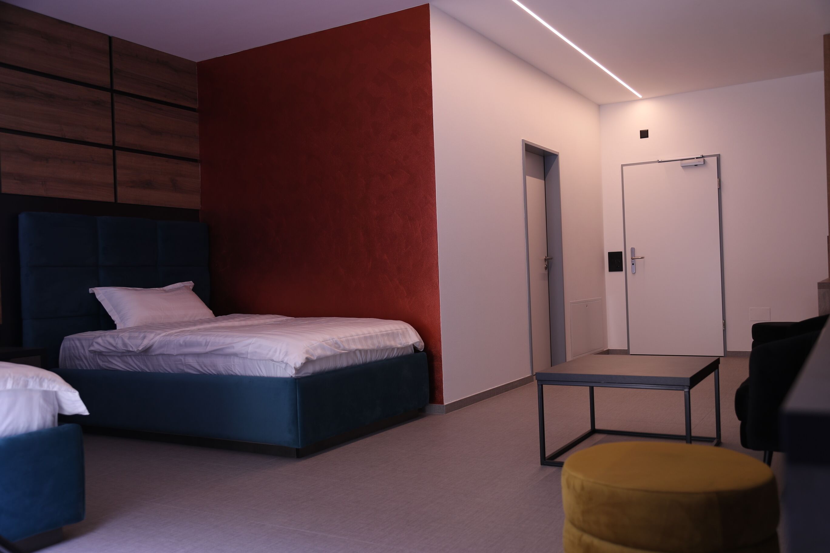A modern hotel room with a bed, a small table, a yellow ottoman, and a door.