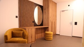 A room with a wooden vanity, a mirror, a yellow ottoman, a yellow chair, and a white door.