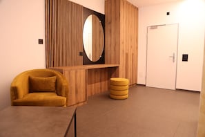 Una habitación con un tocador de madera, un espejo, un reposapiés amarillo, una silla amarilla y una puerta blanca.