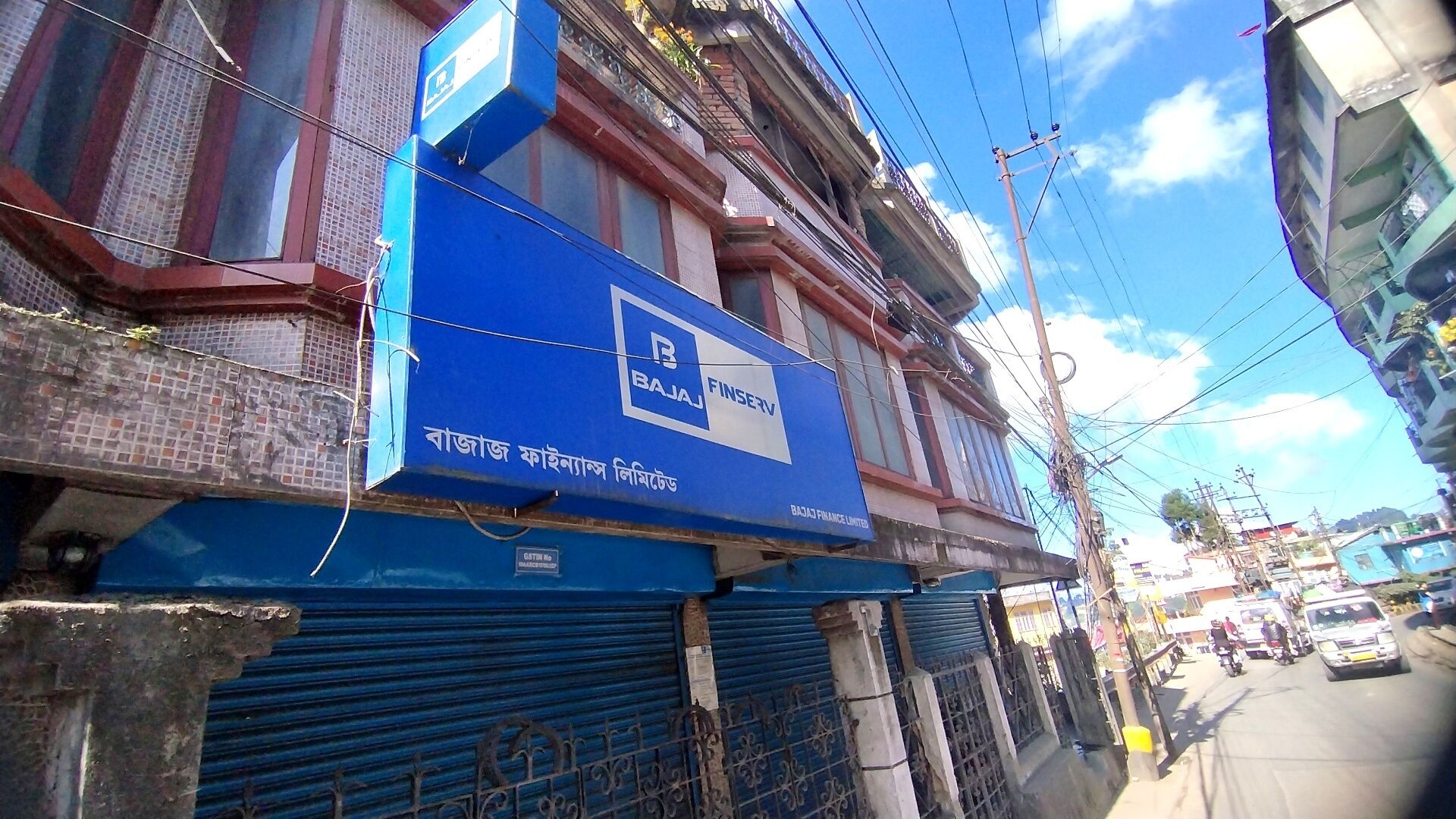 A building with a blue sign for Bajaj Finserv, a closed metal shutter, and a street with vehicles and power lines.