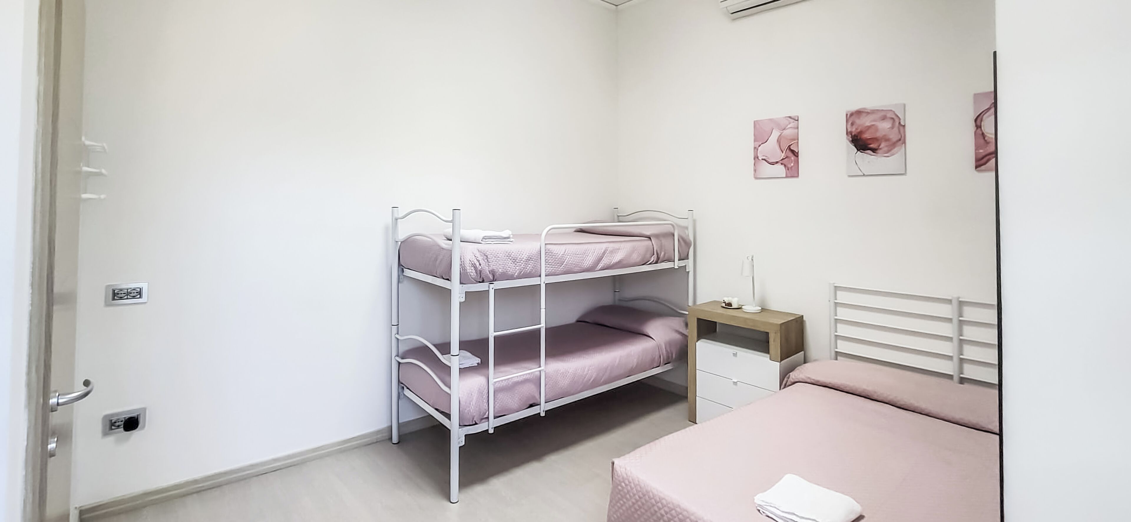 Ein Stockbett mit Schreibtisch und Stuhl, ein Nachttisch mit Lampe sowie drei an der Wand hängende Blumenbilder.