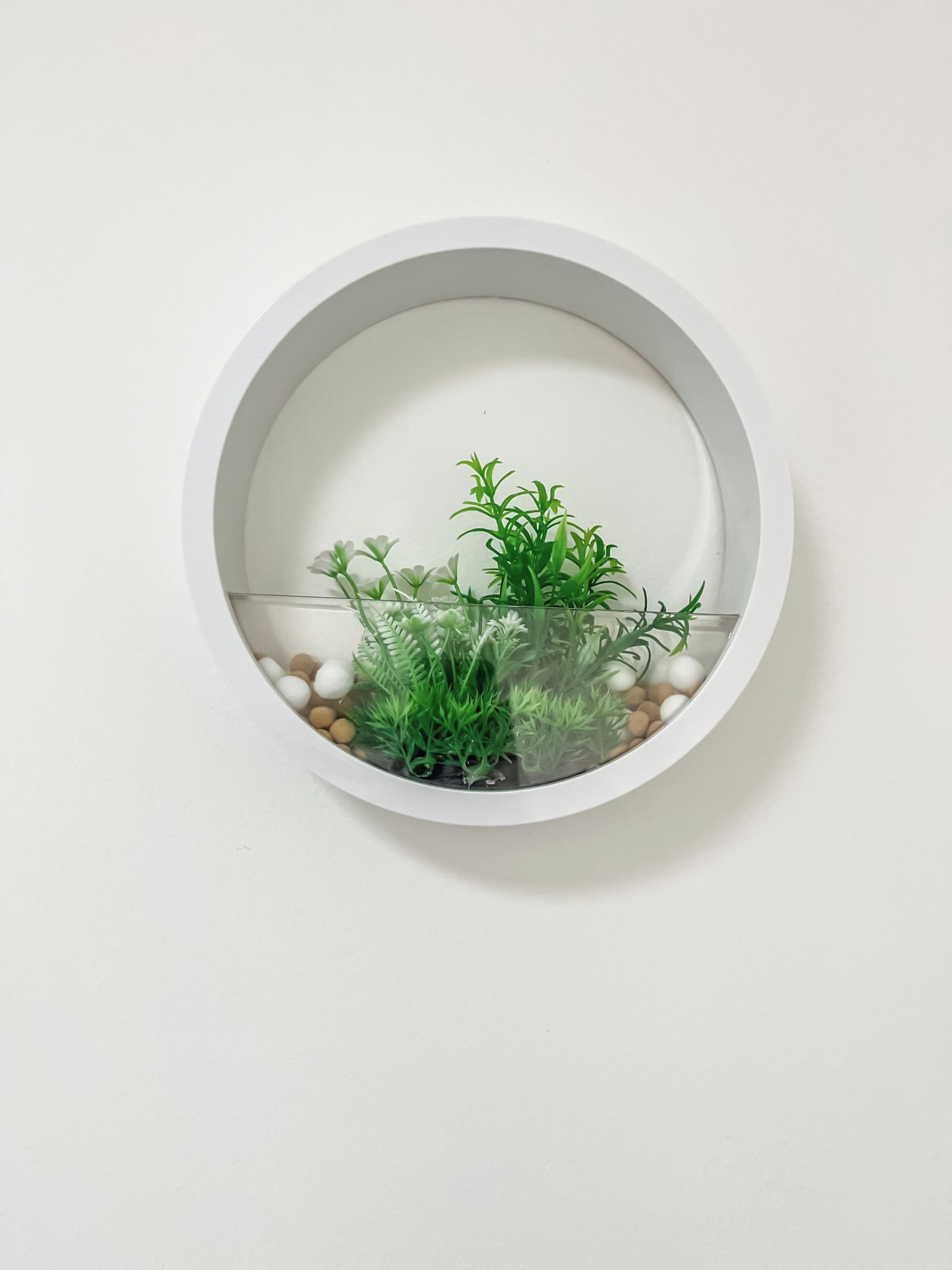 Un acuario redondo blanco fijado a la pared, con plantas acuáticas verdes y guijarros.