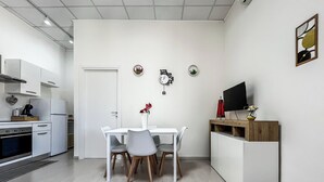 Eine gemütliche Küchenzeile mit weißen Schränken, einem Esstisch mit Stühlen, einem kleinen Fernseher auf einem Holzgestell und einer Uhr an der Wand.