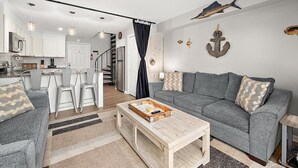 A modern living room with a grey sofa, a coffee table, and a dining area with chairs and a kitchen in the background. - Great Location for Sunday Fireworks and Easy Walk to the Beach! Has a Pool! (Ocean City)