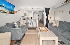 A modern living room with grey sofas, a coffee table, and a TV displaying a beach scene.