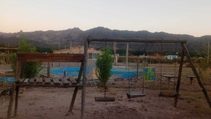 Ein Spielplatz mit Schaukeln und einem kleinen Pool, umgeben von einem befestigten Bereich und Gebäuden im Hintergrund.