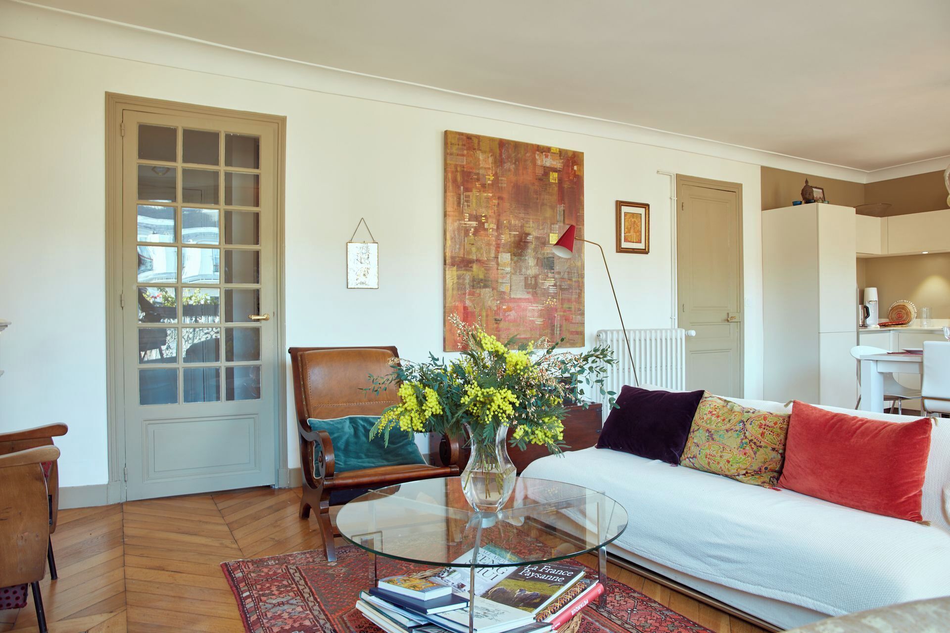 Ein Wohnzimmer mit einer Couch, einem Glastisch, einem Sessel, einem Bücherregal und einem großen abstrakten Gemälde an der Wand.