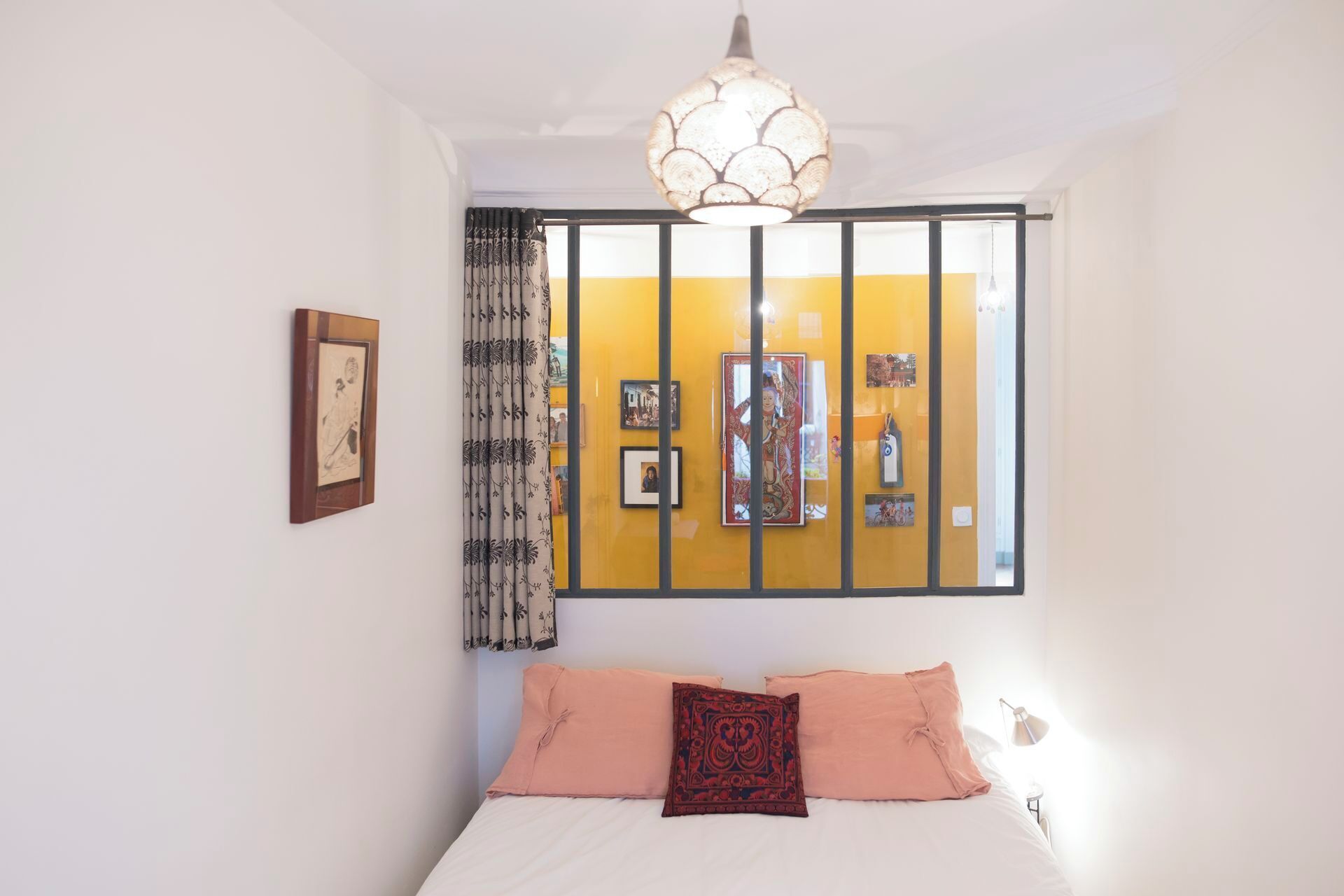 A bedroom with a bed, a hanging light fixture, a window with curtains, and wall-mounted artwork.