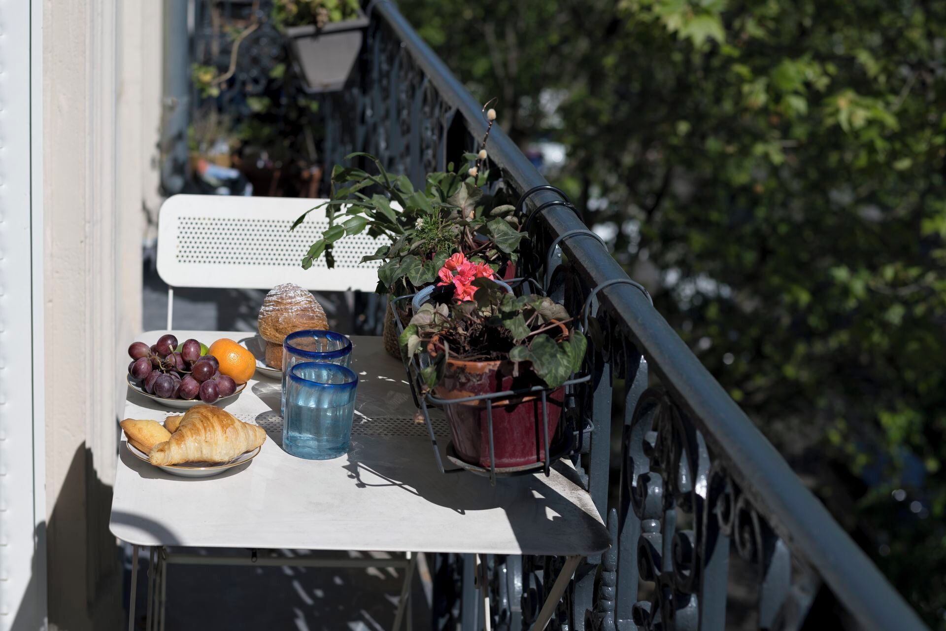 A balcony with a table set with grapes, oranges, croissants, and a potted plant with red flowers.