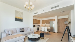 A modern living room with a sofa, coffee table, and ottoman, a dining area with chairs, and a kitchen in the background. - PVH - Executive Residences 2BR (Dubai)