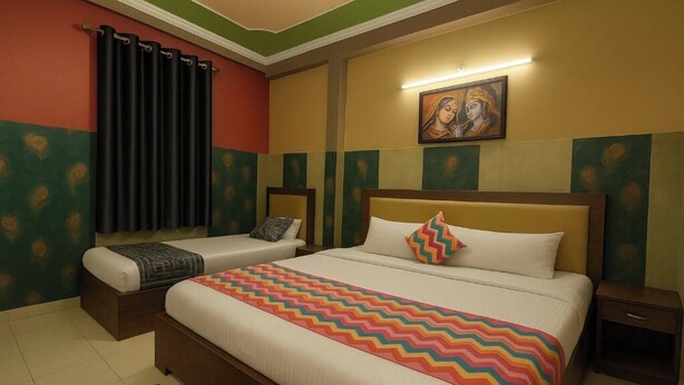 A hotel room with two beds, a nightstand, a wall painting, and a window with curtains. - Hotel Shiri Krishna  (Mathura)