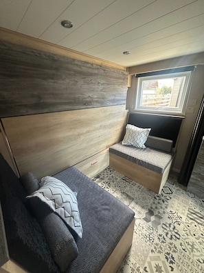 A compact, well-lit room with a bed, a small seating area with cushions, a window, and a patterned floor.