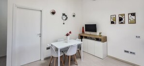 Una habitación compacta con una puerta blanca, una mesa de comedor con sillas, un televisor sobre un mueble de madera y un estante pequeño con adornos.
