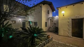A courtyard with a paved walkway, a palm tree, and a building with a stone entrance. - Dolia Court - Dolianova (Dolianova)