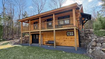 Une cabane en rondins à deux étages, dotée d’un balcon, de fondations en pierre et d’une terrasse en bois.