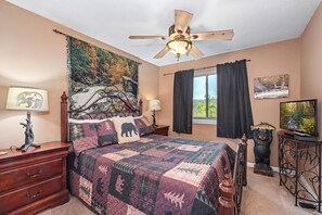 A bedroom with a bed, a ceiling fan, a tapestry, a window with curtains, a nightstand, a dresser, a lamp, and a bear statue. - Escape to the serene beauty of Gatlinburg with Mountain View Memories Unit #2102 (Gatlinburg)
