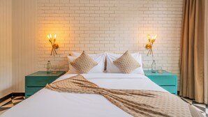 Hotellihuone, jossa on valkoisesta tiilestä tehty seinä, kuviollinen päiväpeite sängyllä, kaksi yöpöytää lampuineen ja turkoosinsininen yöpöytä.