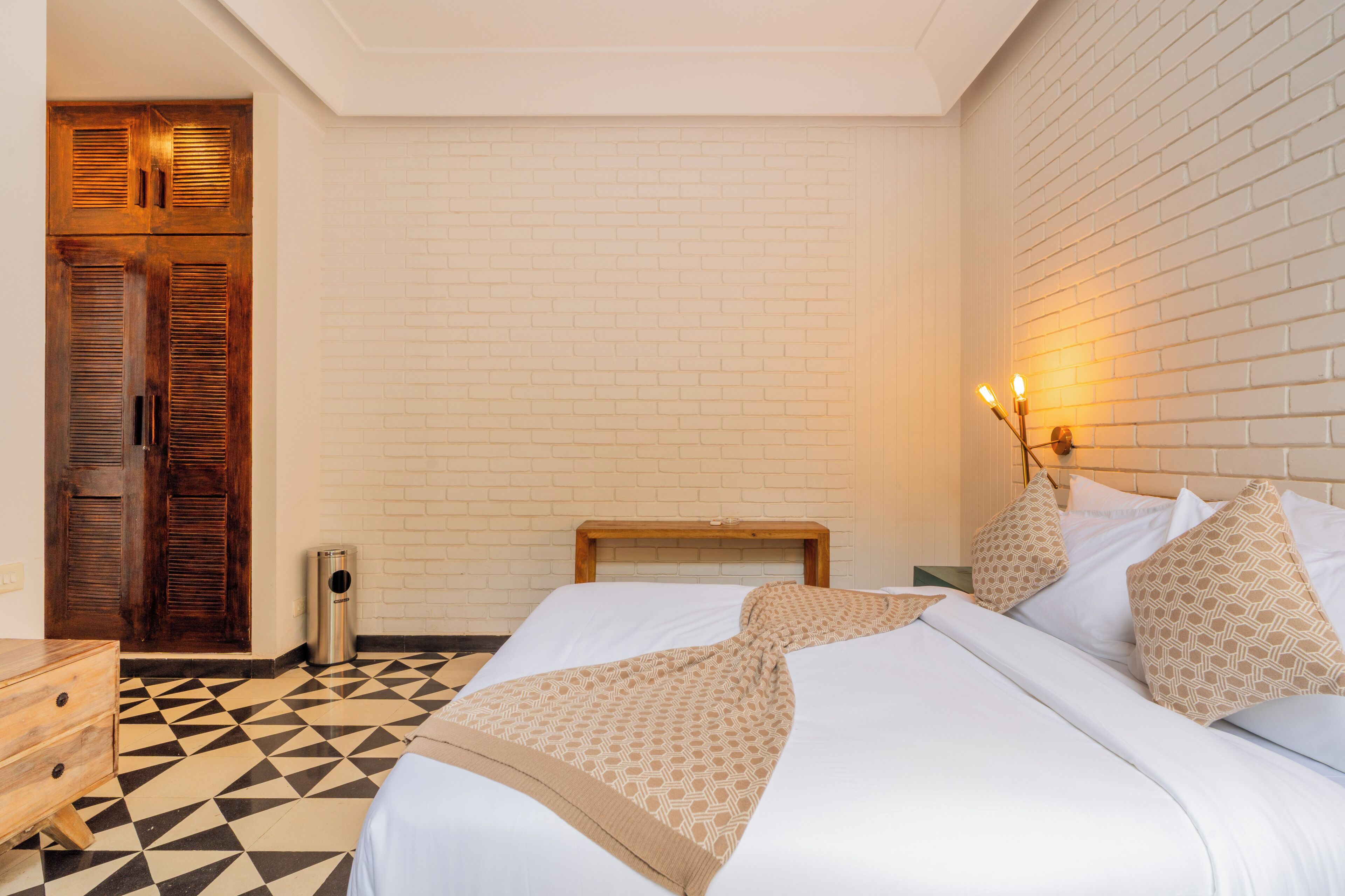 A hotel room with a checkered floor, a bed with patterned bedding, a wooden headboard, and a brick wall.