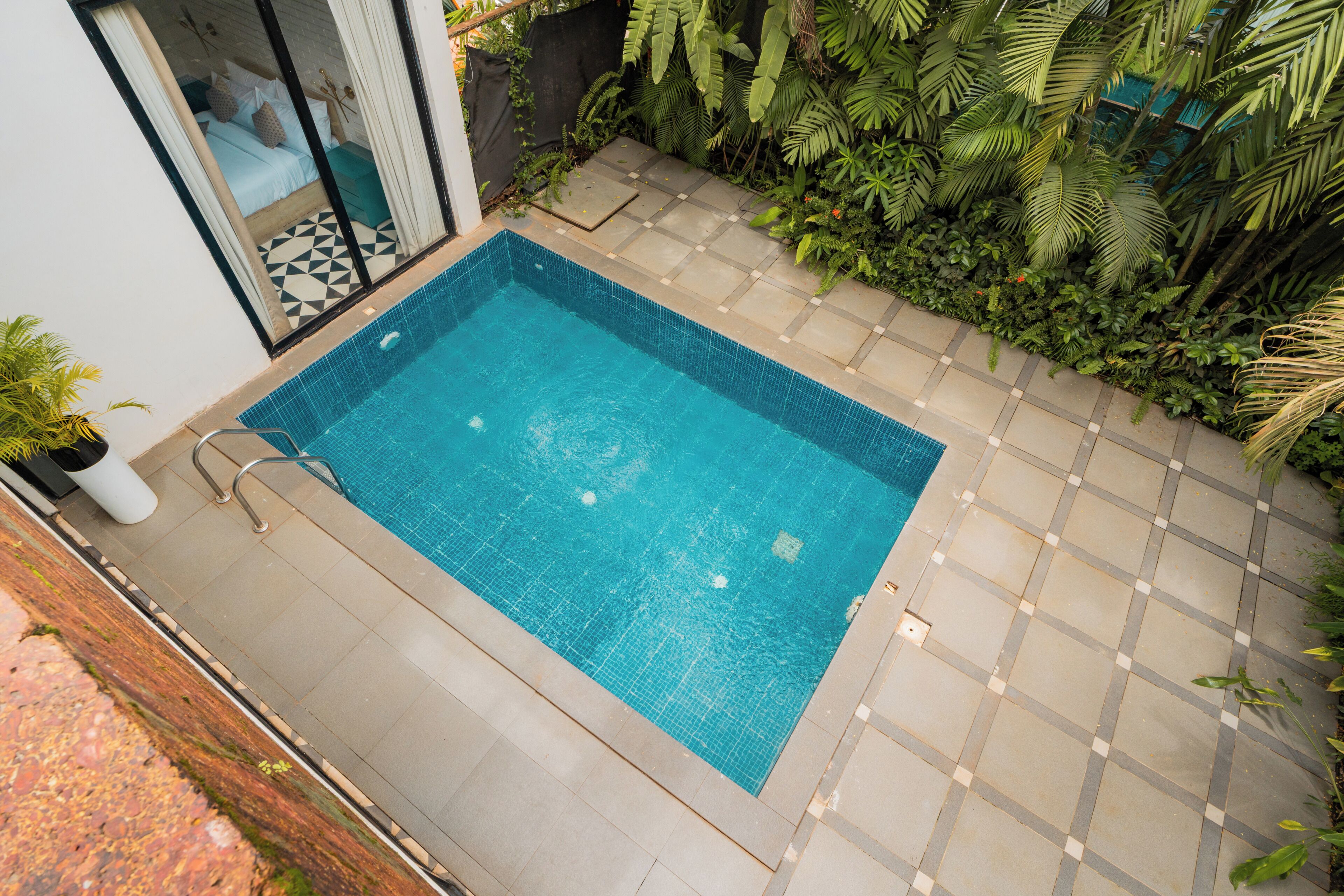 Ein rechteckiger Pool mit Fliesen, umgeben von einer gefliesten Terrasse und üppiger Bepflanzung.