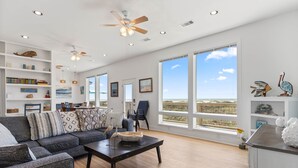 A spacious living room with a large window, a ceiling fan, and a bookshelf filled with books and decorative items. - Driftwood Beach House: Your Perfect Getaway in Matagorda (Matagorda)
