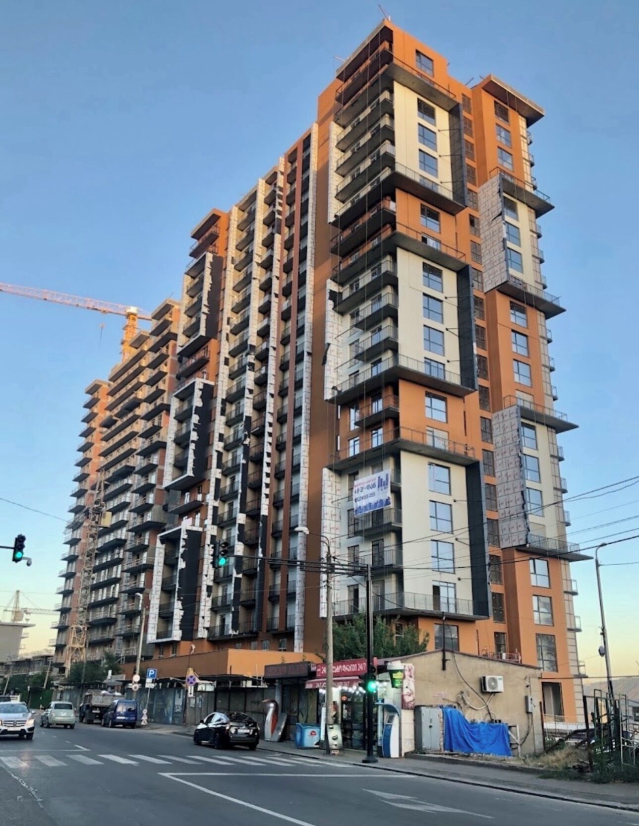 Un immeuble résidentiel à plusieurs étages, doté de balcons, sur lequel on peut voir une grue de construction, et une rue avec des voitures et des feux de signalisation.