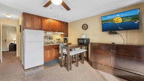 A compact kitchen with wooden cabinets, a refrigerator, a microwave, and a dining table with stools. A flat-screen TV is mounted on the wall above the counter.