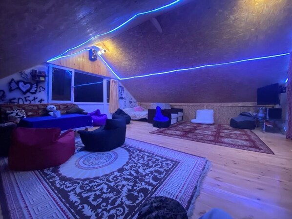 Une pièce mansardée avec un plancher en bois, un tapis à motifs, des fauteuils en forme de poire et un plafond éclairé d’une lumière bleue.