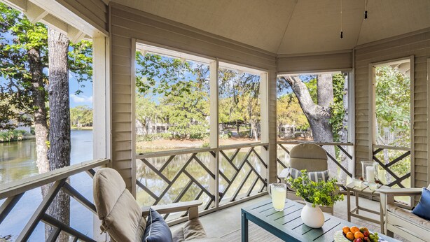 A covered outdoor seating area with a view of a lake, a table with a pitcher and fruit, and a chair with a cushion. - Stunning remodeled Turtle Cove (Kiawah Island)