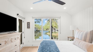 A bedroom with a bed, a TV, a dresser, a window with a view of a lake, and a ceiling fan. - Stunning remodeled Turtle Cove (Kiawah Island)