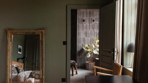 A room with a large ornate mirror, a bed with patterned bedding, a small stool, a wicker basket, and a table with a chair. - Moulin Moulin (Dannes)
