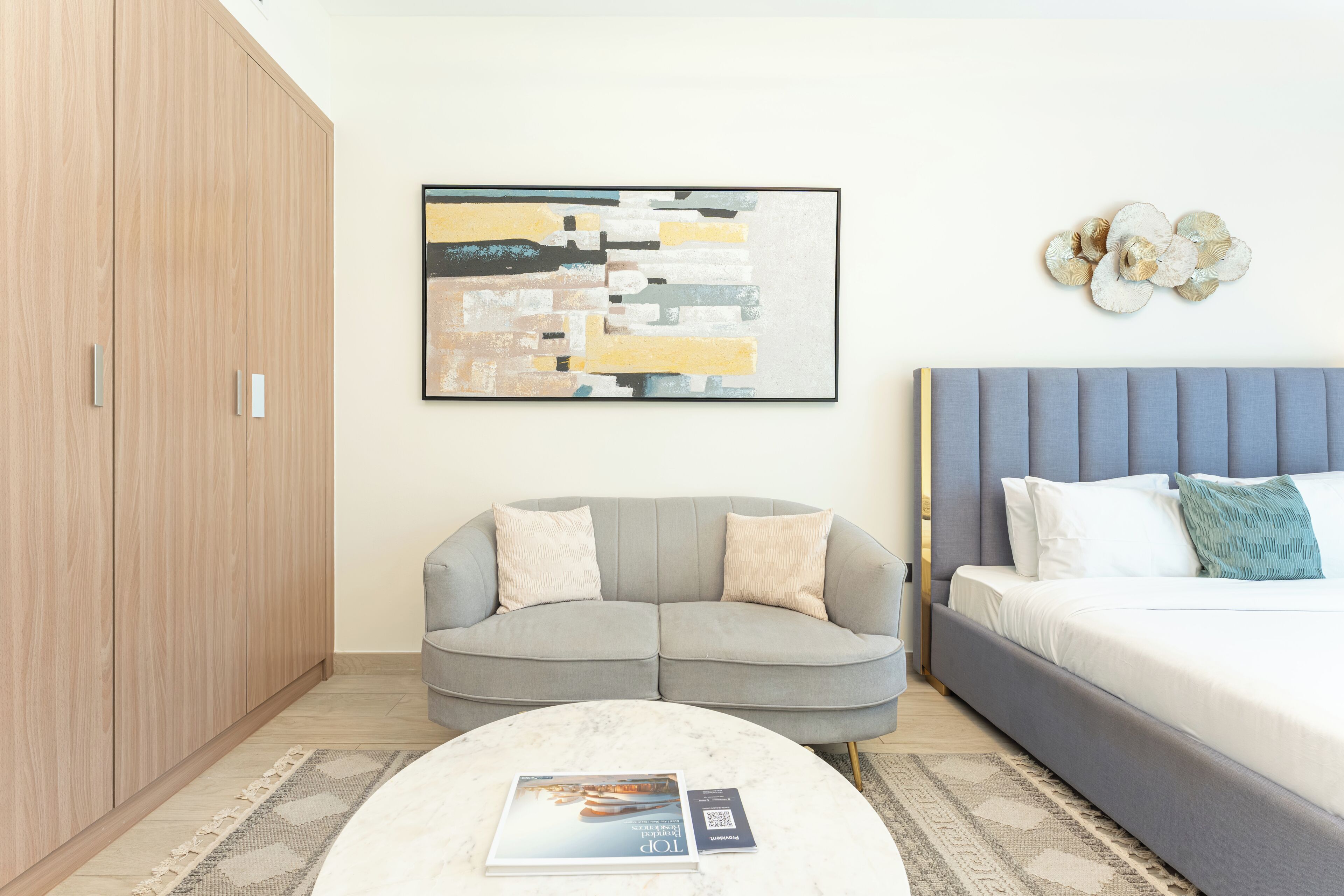 Une chambre à coucher moderne avec un lit, un canapé gris, une armoire en bois et un tableau abstrait encadré.