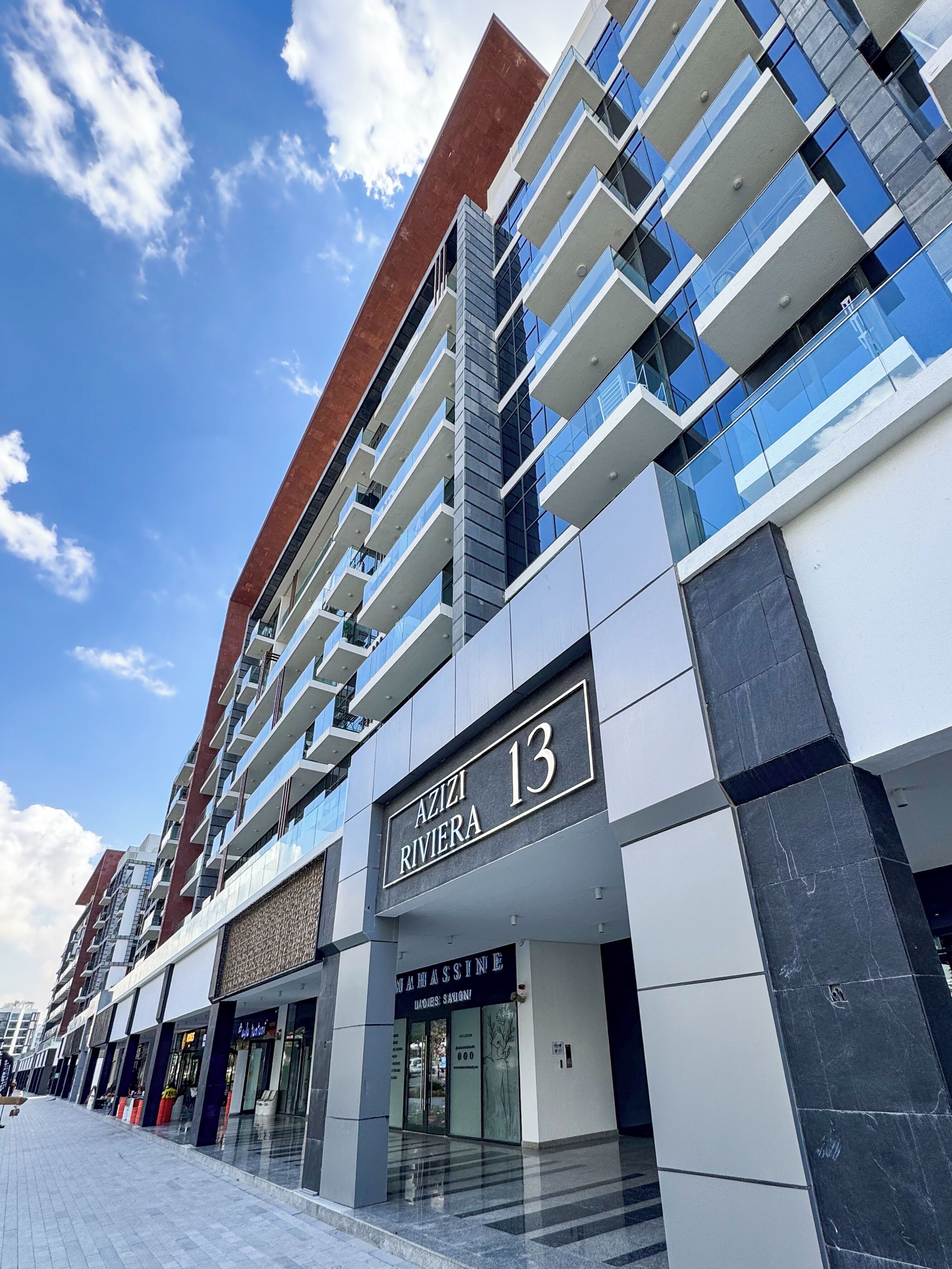 Un edificio moderno con un letrero que dice 'AZIZI RIVIERA 13' y una entrada visible a un establecimiento comercial.