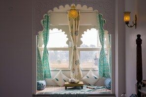 Een kamer met raam, gordijnen, een bed, kussens, een tafel met theepot en een wandlamp.