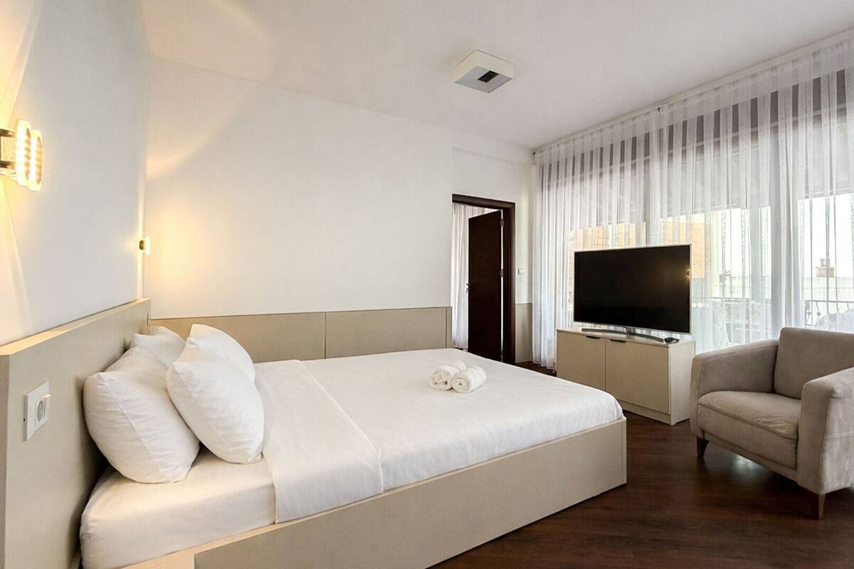 A hotel room with a bed, a TV, a chair, and a large window with curtains.
