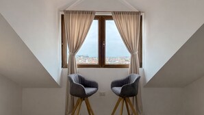 A room with a sloped ceiling, two chairs, a window with curtains, and a city view. - Makadam Hotel (Prizren)