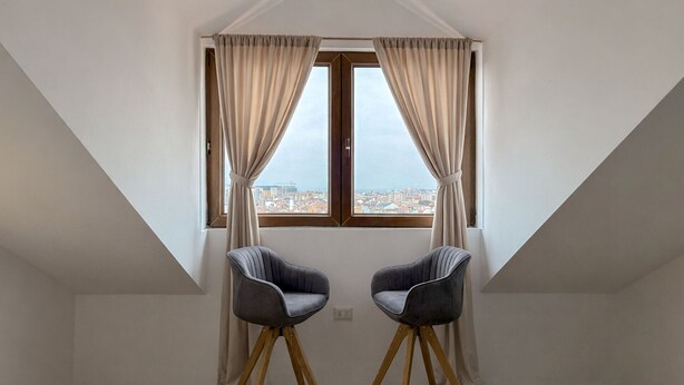Ein Zimmer mit schrägen Wänden, zwei Stühlen, einem fenster mit Vorhängen und Blick auf die Stadt.