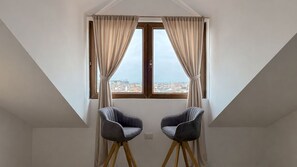 Ein Zimmer mit schrägen Wänden, zwei Stühlen, einem fenster mit Vorhängen und Blick auf die Stadt.
