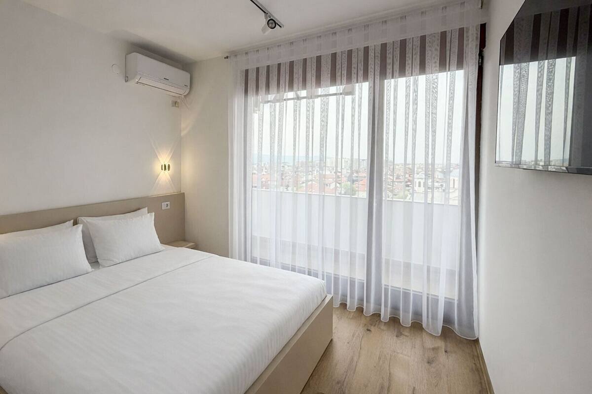 A hotel room with a bed, two wall-mounted lights, an air conditioning unit, and a large window with sheer curtains.