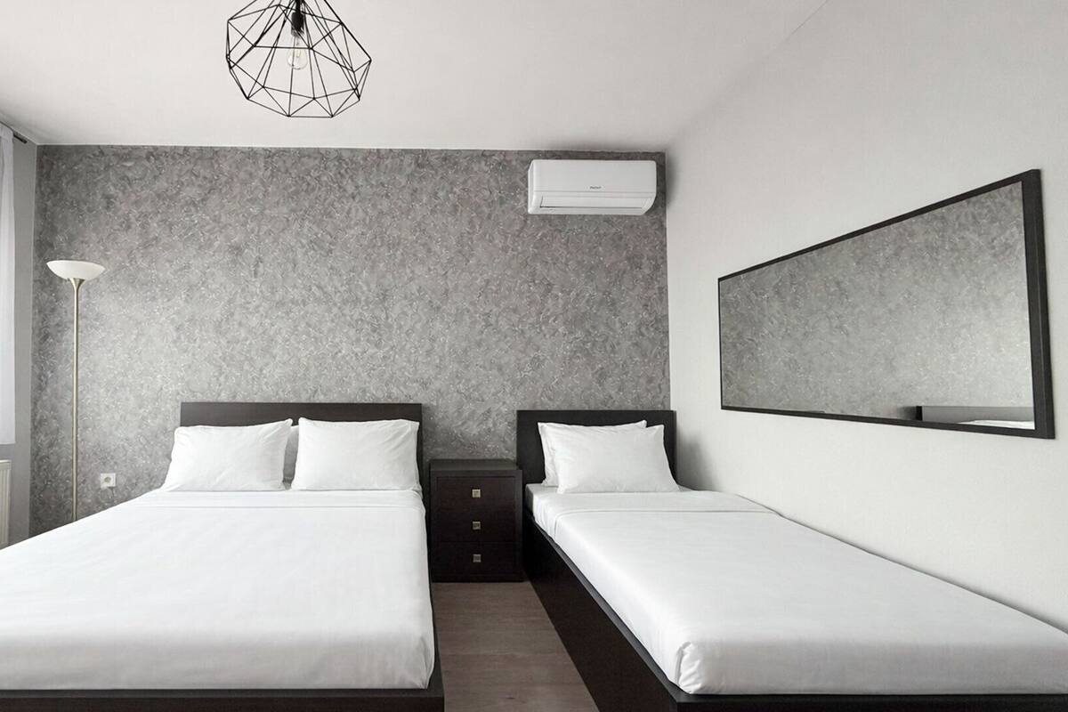 A modern hotel room with two single beds, a large mirror, a geometric light fixture, and a textured gray wall.